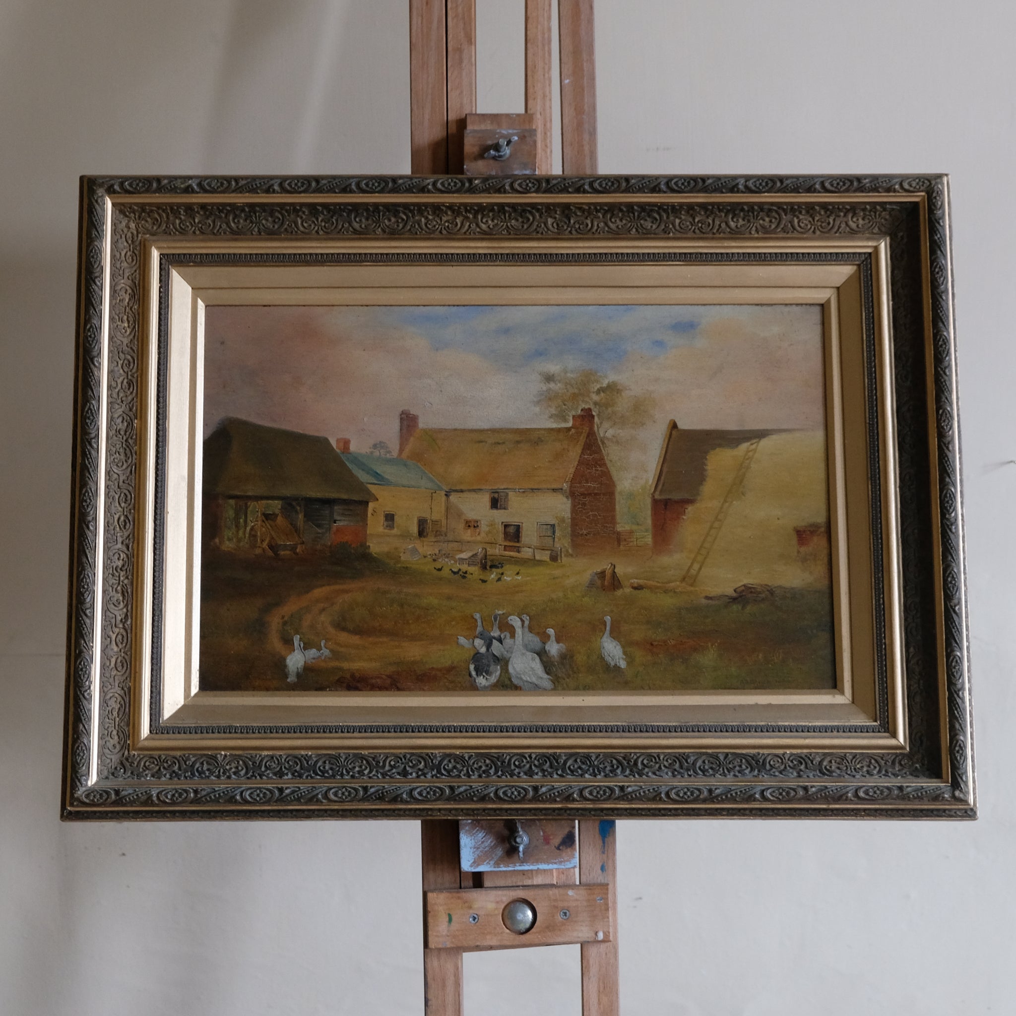 Framed painting of a rural scene with buildings and animals on an easel.