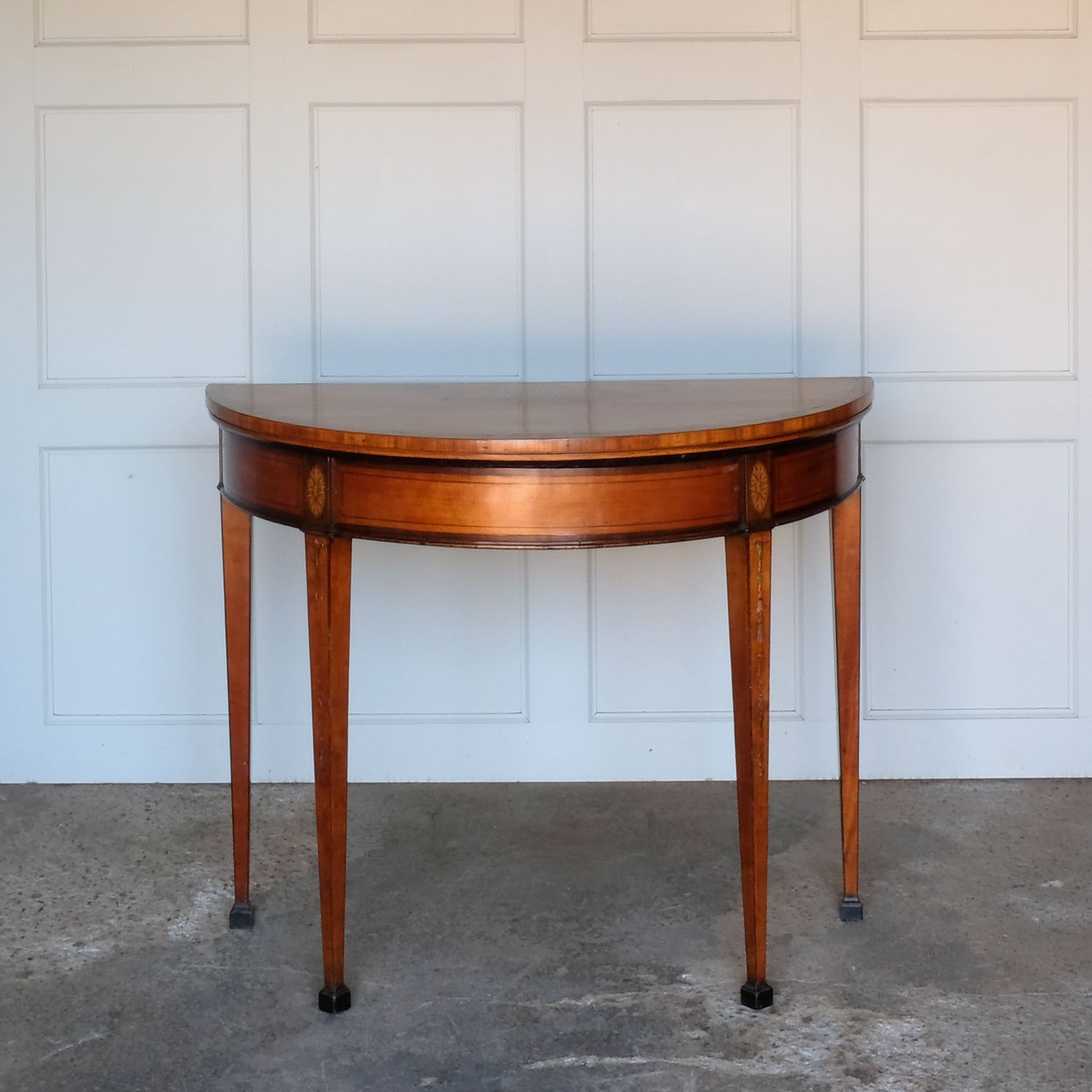 A George III satinwood and marquetry demilune folding card table, with a fold over top fitted with green baize. The top features a beautiful fan inlay and crossbanding around the outside, with the frieze incorporating purple heart crossbanding, over four elegantly tapering legs with floral painted decoration. The rear legs are hinged to swing out and support the folding top. In very good, sturdy overall condition. Would work well as a side console table, or a centre hall table.