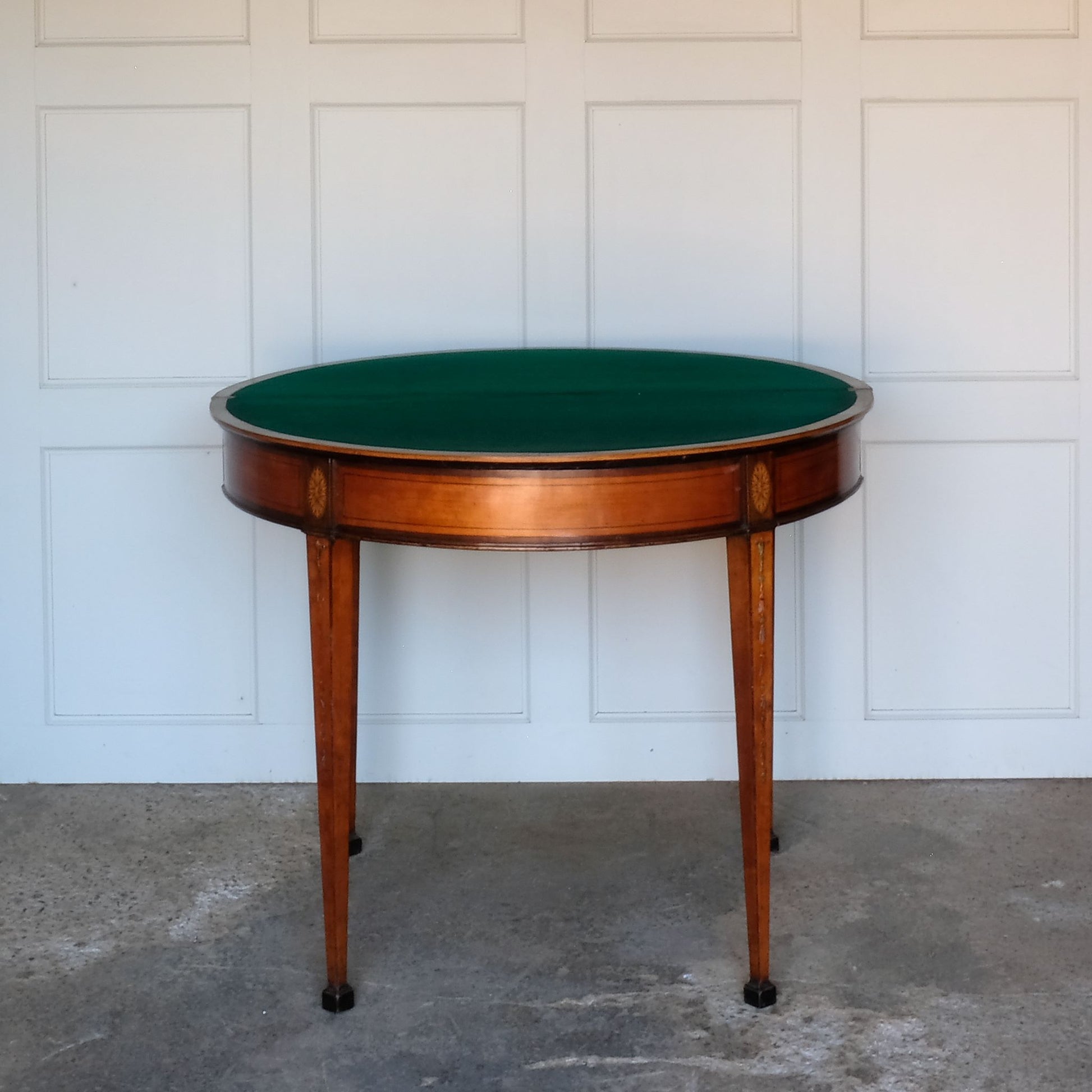 A George III satinwood and marquetry demilune folding card table, with a fold over top fitted with green baize. The top features a beautiful fan inlay and crossbanding around the outside, with the frieze incorporating purple heart crossbanding, over four elegantly tapering legs with floral painted decoration. The rear legs are hinged to swing out and support the folding top. In very good, sturdy overall condition. Would work well as a side console table, or a centre hall table.