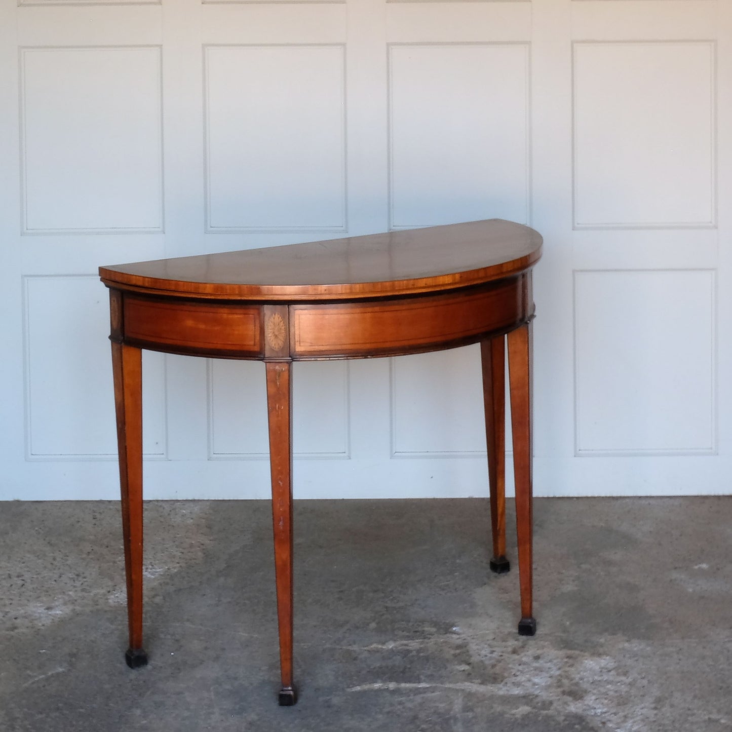 A George III satinwood and marquetry demilune folding card table, with a fold over top fitted with green baize. The top features a beautiful fan inlay and crossbanding around the outside, with the frieze incorporating purple heart crossbanding, over four elegantly tapering legs with floral painted decoration. The rear legs are hinged to swing out and support the folding top. In very good, sturdy overall condition. Would work well as a side console table, or a centre hall table.