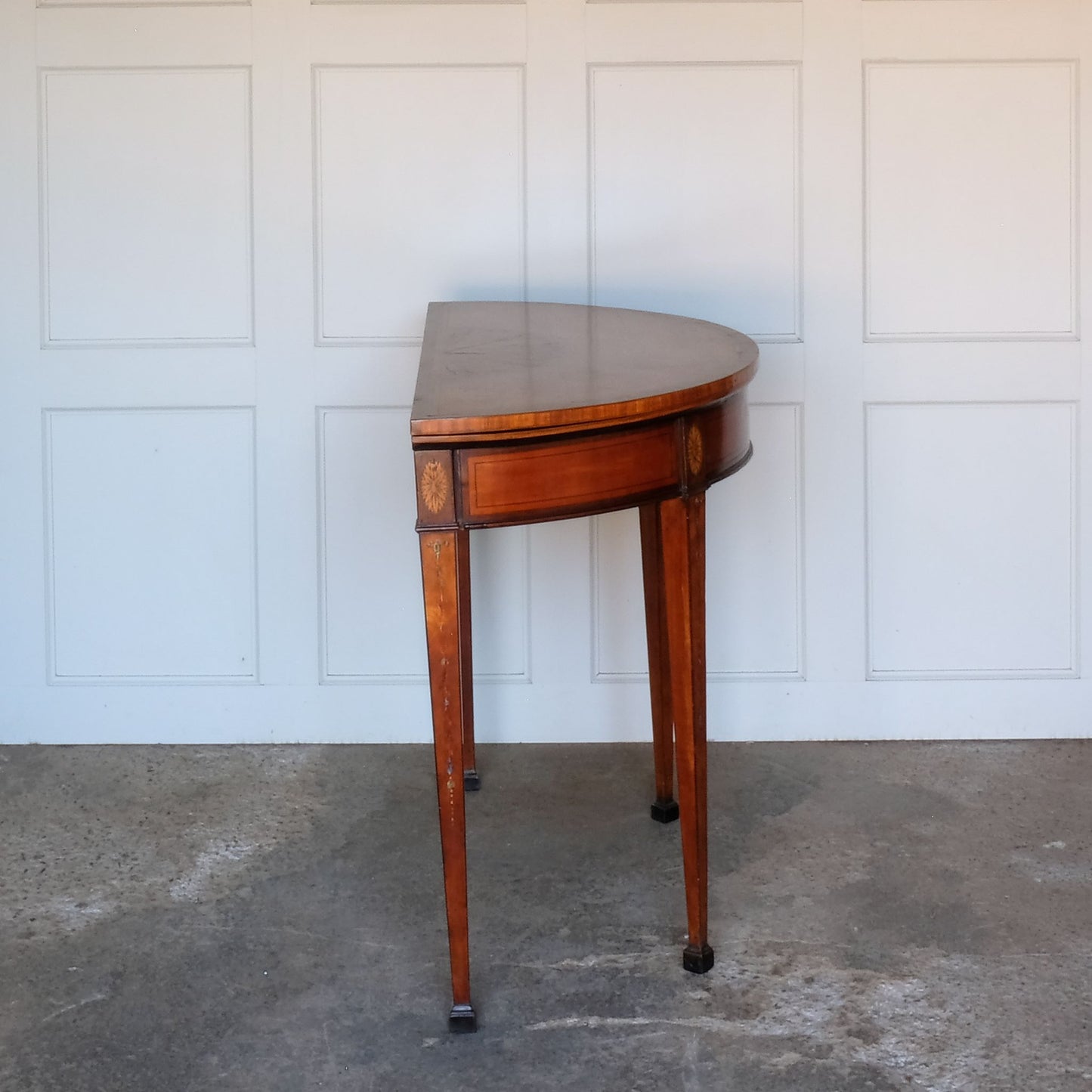 A George III satinwood and marquetry demilune folding card table, with a fold over top fitted with green baize. The top features a beautiful fan inlay and crossbanding around the outside, with the frieze incorporating purple heart crossbanding, over four elegantly tapering legs with floral painted decoration. The rear legs are hinged to swing out and support the folding top. In very good, sturdy overall condition. Would work well as a side console table, or a centre hall table.