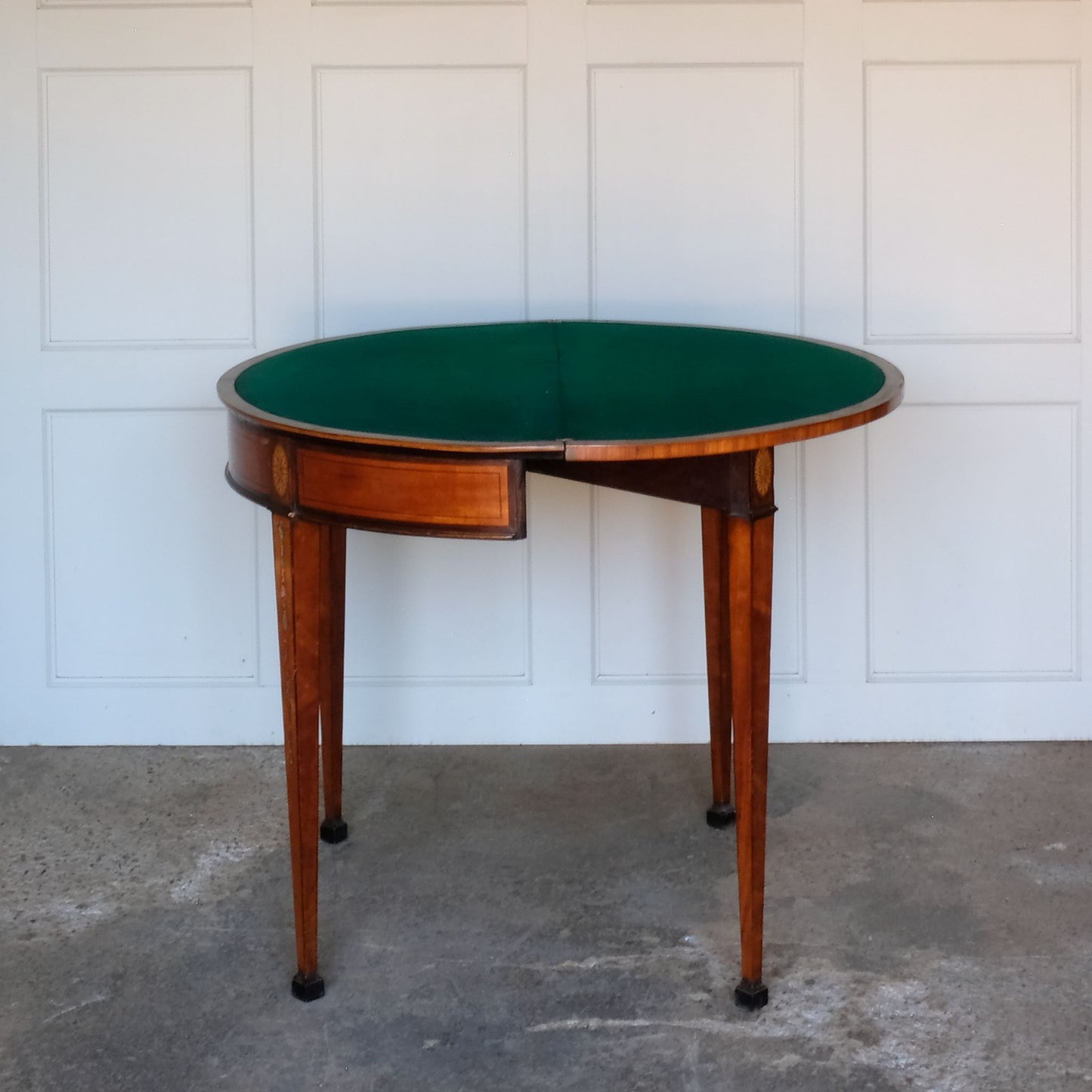 A George III satinwood and marquetry demilune folding card table, with a fold over top fitted with green baize. The top features a beautiful fan inlay and crossbanding around the outside, with the frieze incorporating purple heart crossbanding, over four elegantly tapering legs with floral painted decoration. The rear legs are hinged to swing out and support the folding top. In very good, sturdy overall condition. Would work well as a side console table, or a centre hall table.