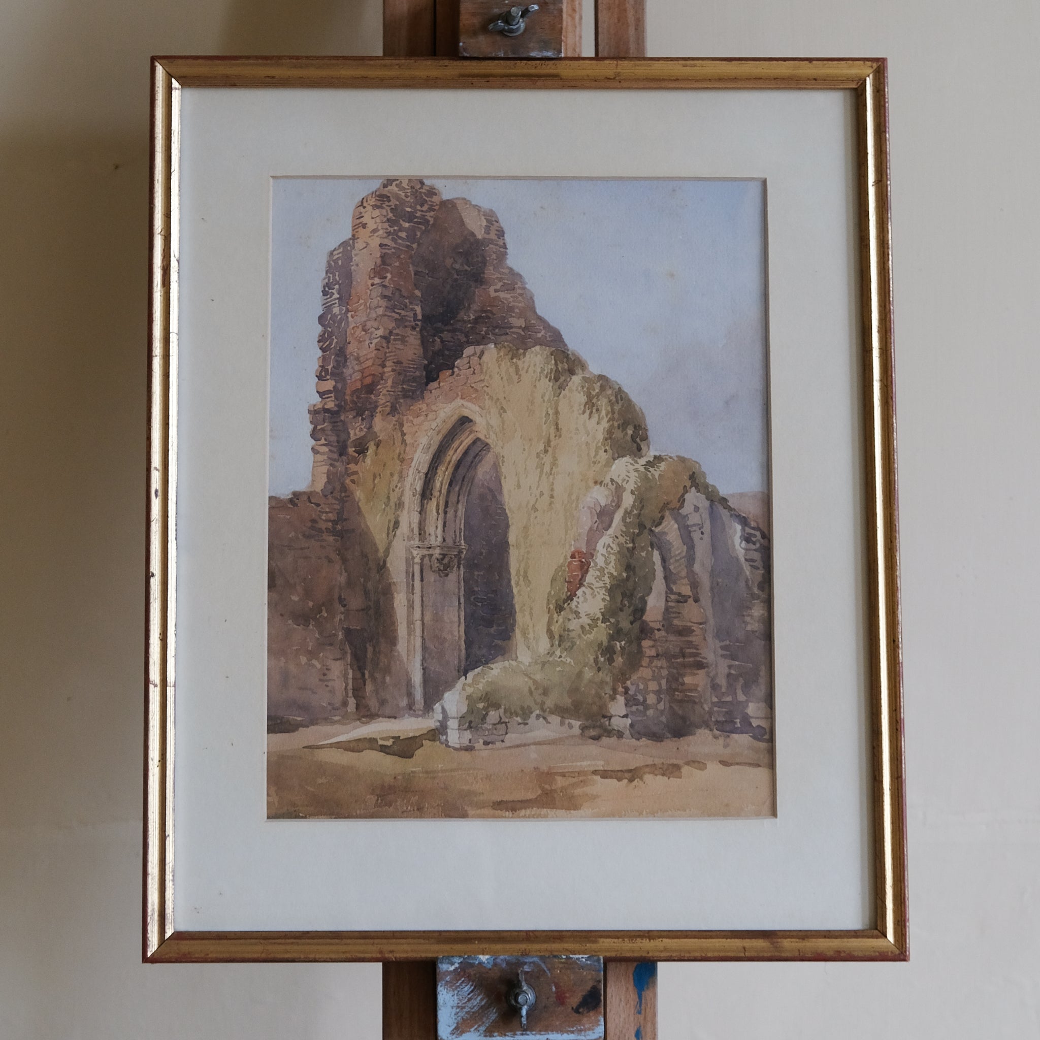 Framed artwork of a ruins scene on an easel against a plain wall.