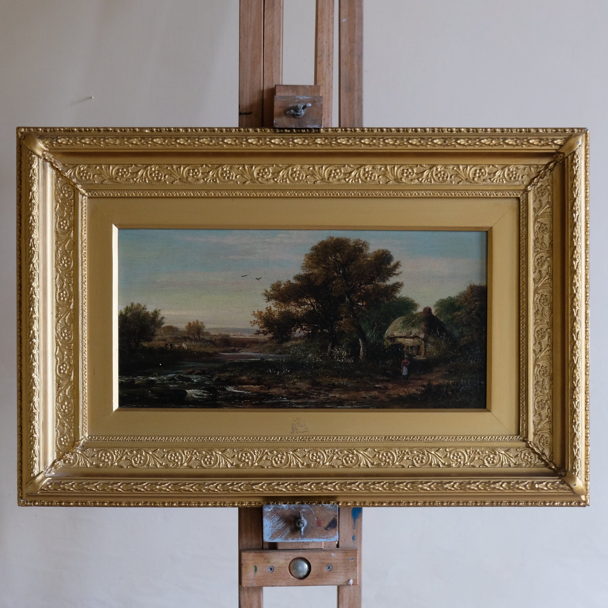 Painting of a landscape with trees and a person on an easel with a gold frame.