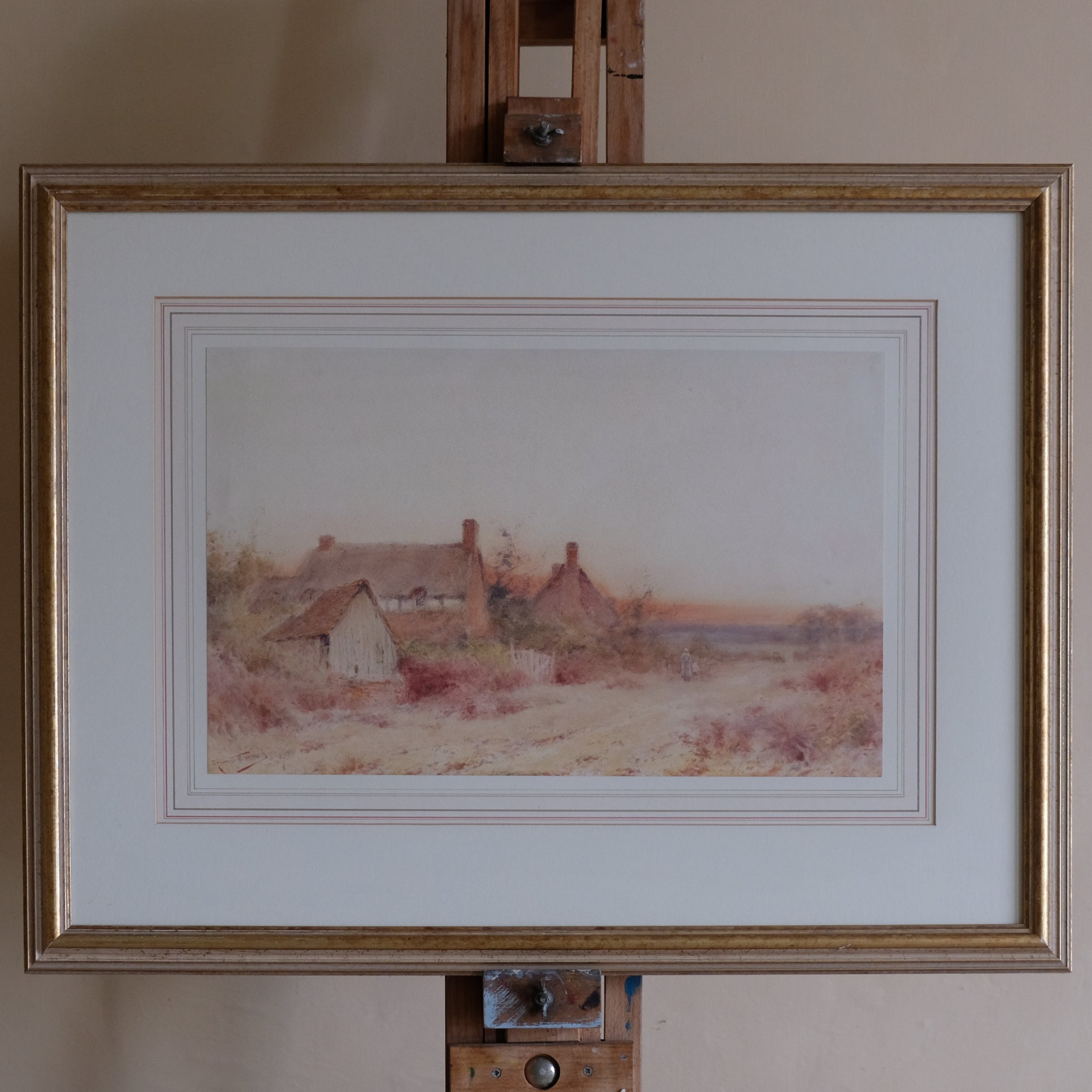 Framed landscape painting of a village scene on an easel.