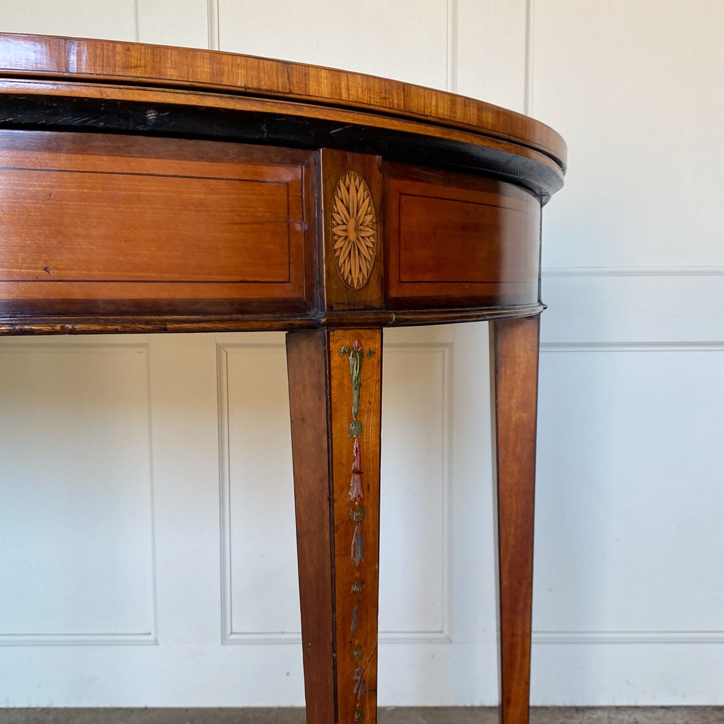 A George III satinwood and marquetry demilune folding card table, with a fold over top fitted with green baize. The top features a beautiful fan inlay and crossbanding around the outside, with the frieze incorporating purple heart crossbanding, over four elegantly tapering legs with floral painted decoration. The rear legs are hinged to swing out and support the folding top. In very good, sturdy overall condition. Would work well as a side console table, or a centre hall table.