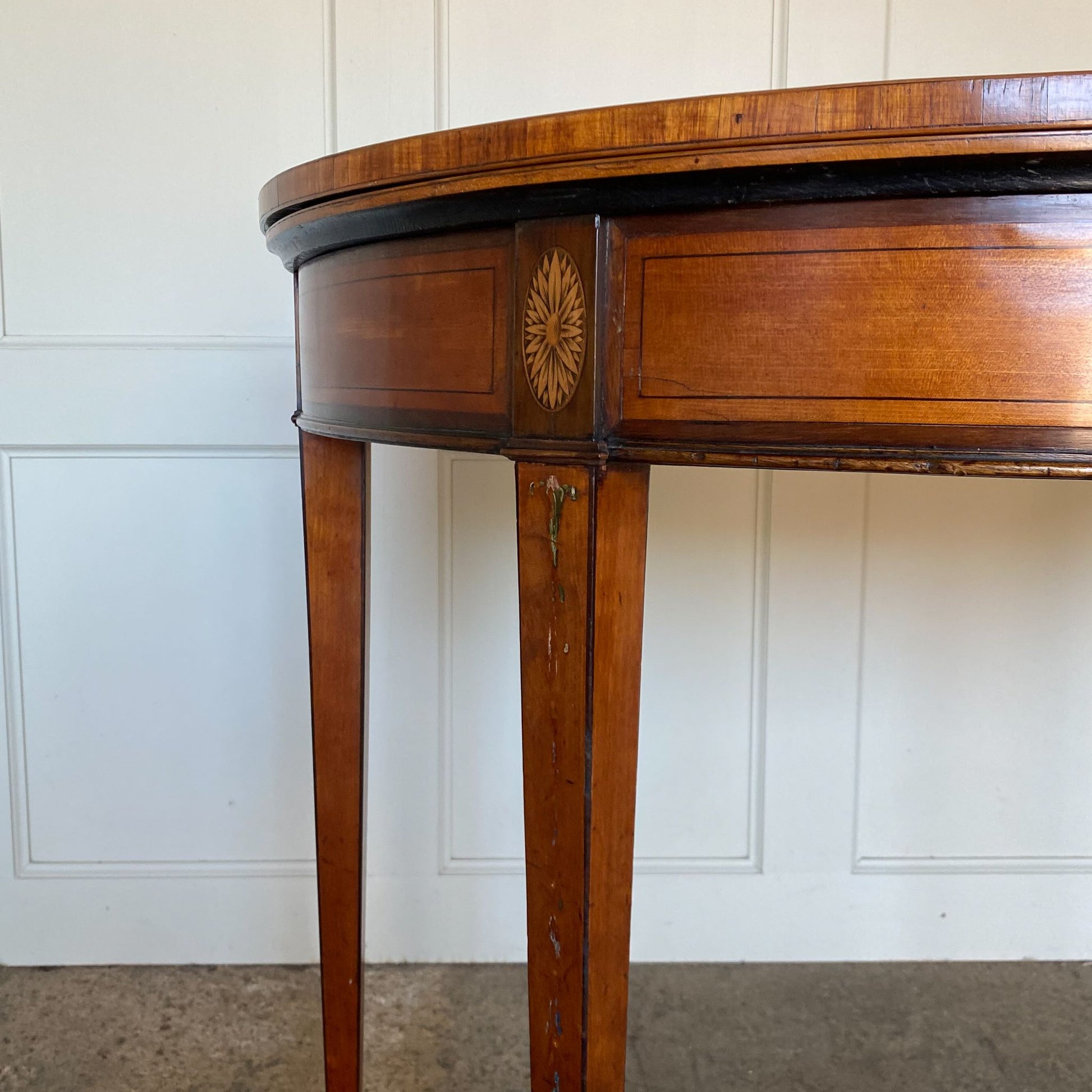 A George III satinwood and marquetry demilune folding card table, with a fold over top fitted with green baize. The top features a beautiful fan inlay and crossbanding around the outside, with the frieze incorporating purple heart crossbanding, over four elegantly tapering legs with floral painted decoration. The rear legs are hinged to swing out and support the folding top. In very good, sturdy overall condition. Would work well as a side console table, or a centre hall table.