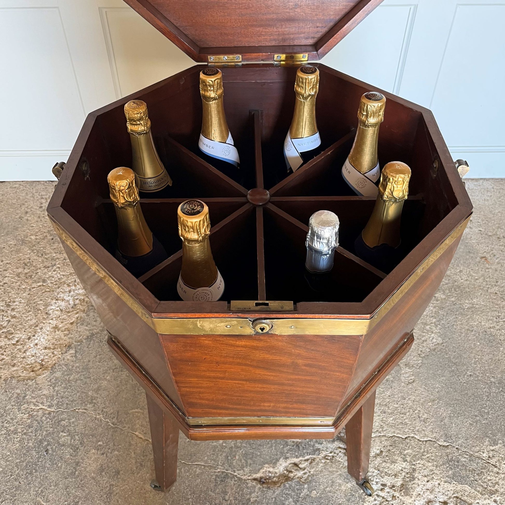 A George III mahogany octagonal brass bound wine cooler on its original stand. The hinged lid encloses an eight division interior, with brass carrying handles on the side and a fixed stand with square tapering legs on brass castors. A beautiful patina throughout. A small piece of brass trim is missing around the face of the lock, the catch of which does not completely close, leaving a gap at the front of c. 0.5cm when shut, as reflected in the price.